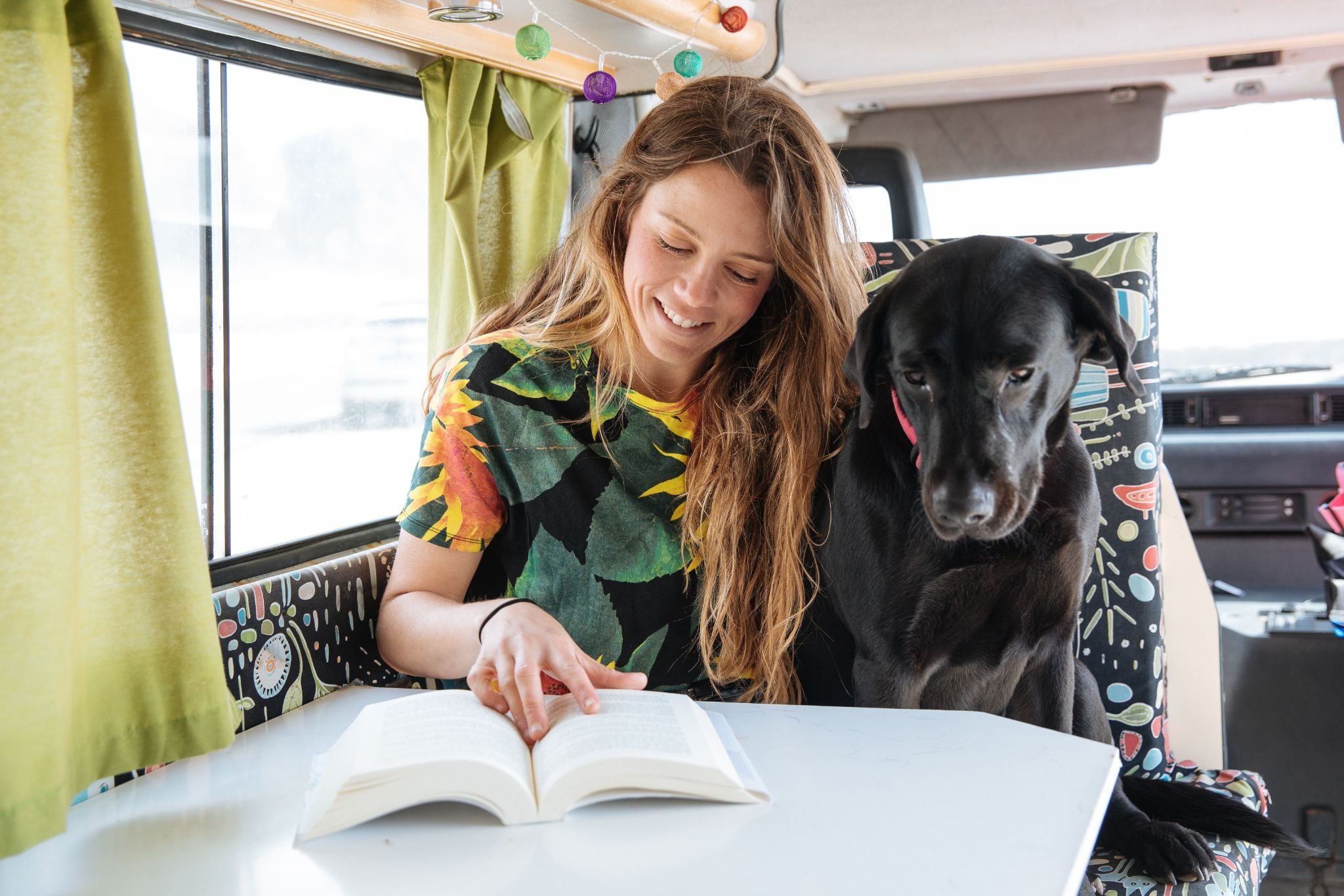 Woman reading a book indoors with a black dog sitting beside her, illustrating companionship and the emotional benefits of having a dog